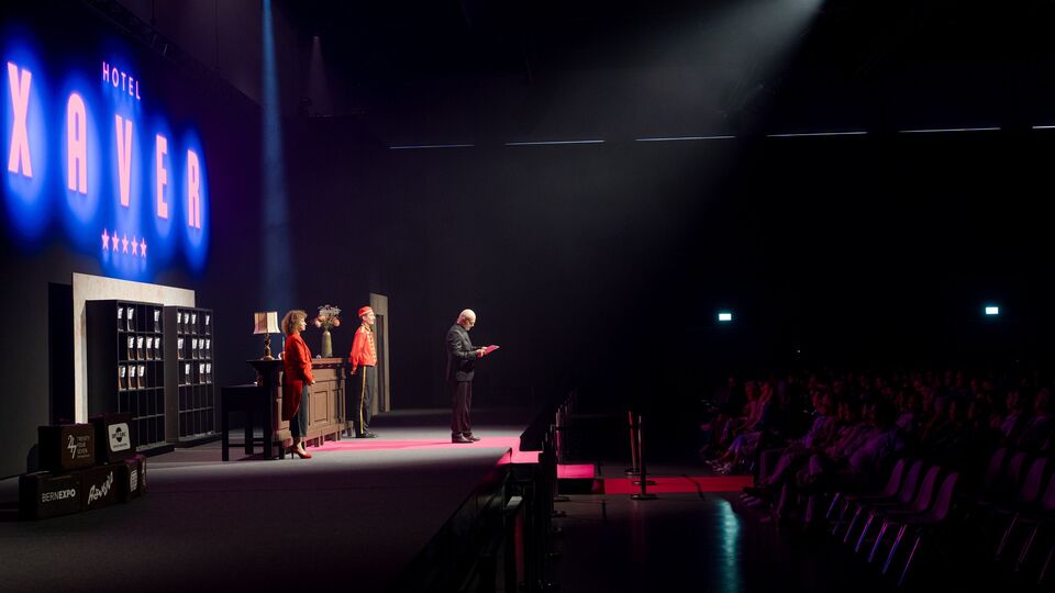 XAVER Award 2026 in der Festhalle Bern: Hotelkulisse mit Leuchtschrift «Hotel XAVER», Moderation und Publikum.