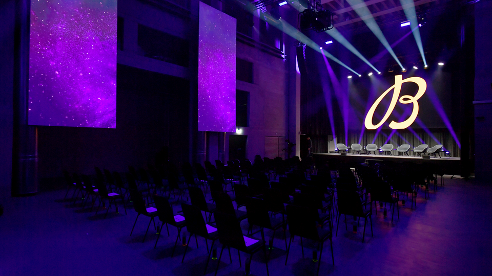 Die mit stimmungsvoller Lichttechnik für eine Produktlancierung in violettes Licht eingetauchte Eventhalle, auf der Bühne ein grosses Logo des zu präsentierenden Produkts.