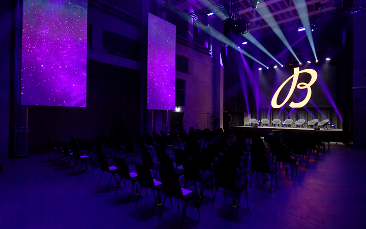 Die mit stimmungsvoller Lichttechnik für eine Produktlancierung in violettes Licht eingetauchte Eventhalle, auf der Bühne ein grosses Logo des zu präsentierenden Produkts.