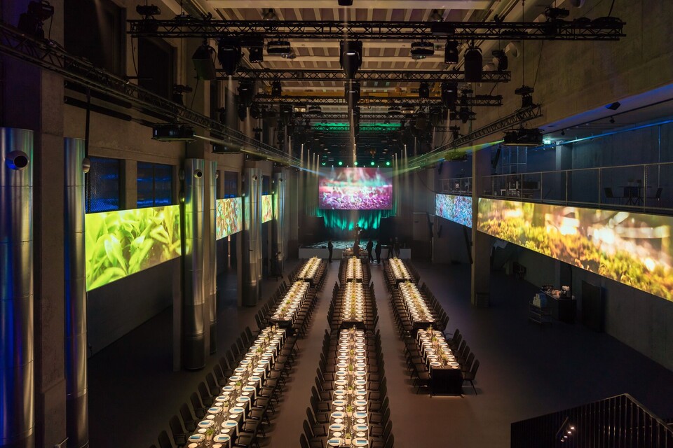 Die für ein grosses Dinner hergerichtete Eventhalle mit hell beleuchteten Tischen und Bildern an den Balustraden der Gallerie.