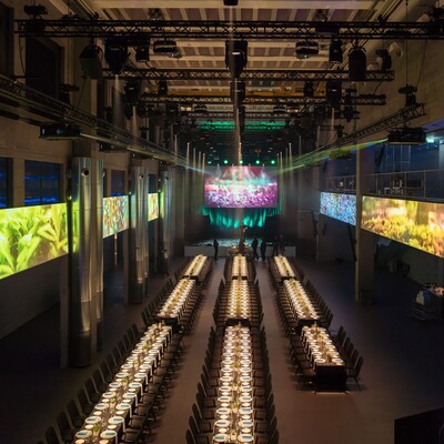 Die für ein grosses Dinner hergerichtete Eventhalle mit hell beleuchteten Tischen und Bildern an den Balustraden der Gallerie.